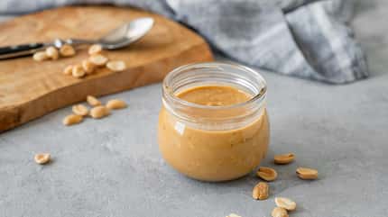 Glass jar filled with creamy peanut butter on grey background with scattered roasted nuts