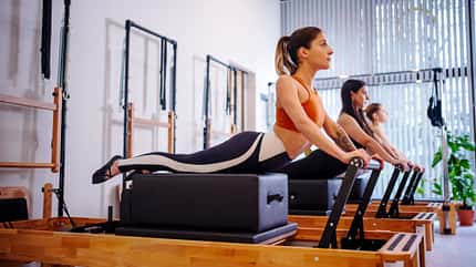 Mehrere Frauen hintereinander in einer Reihe, wie sie an Reformer-Pilates-Maschinen trainieren Mehrere Frauen hintereinander in einer Reihe, wie sie an Reformer-Pilates-Maschinen trainieren