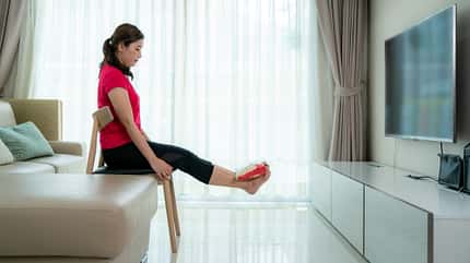 Eine asiatische Frau sitzt auf einem Stuhl und benutzt eine Lebensmittelpackung als Fußgewicht Eine asiatische Frau sitzt auf einem Stuhl und benutzt eine Lebensmittelpackung als Fußgewicht