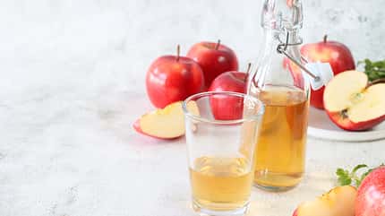 Image of open, swing stopper bottle containing apple cider vinegar by drinking glass, surrounded by red Honeycrisp apples (Malus domestica), whole, halved and sliced fruit, white background, health and wellbeing concept, copy space
