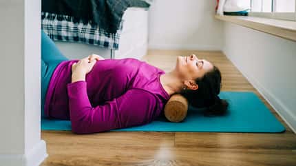 Eine Frau liegt auf einer Yogamatte mit einer Faszienrolle unter dem Nacken