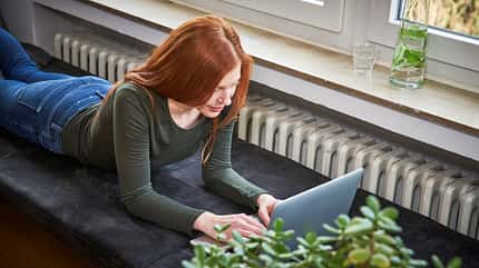 Eine rothaarige Frau liegt auf dem Sofa und arbeitet am Laptop
