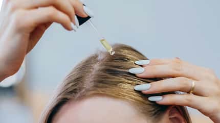 Closeup der Kopfhaut einer Frau, die eine Pipette mit Öl über ihren Haaransatz hält.