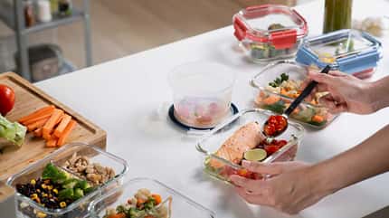 Hände, die eine Brotdose aus Glas mit Essen befüllen. Die Arbeitsfläche ist voll mit frischem Essen und Brotdosen.