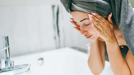 Übers Waschbecken gebeugte Frau, Haare in Handtuch gewickelt und Hände sanft im Gesicht