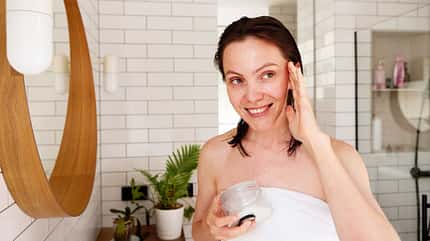 Frau steht lächelnd im Bad und trägt Creme im Gesicht auf Frau steht lächelnd im Bad und trägt Creme im Gesicht auf