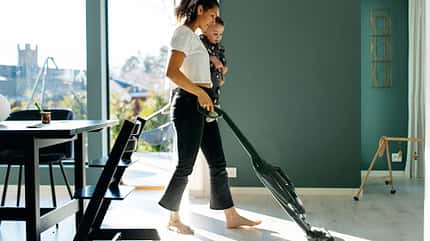 Frau saugt das Wohnzimmer und hält gleichzeitig ihren Sohn im Arm