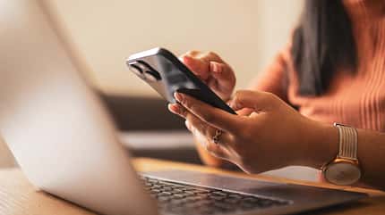 Closeup von Händen einer Frau, die ein Handy halten, im Vordergrund ein Laptop