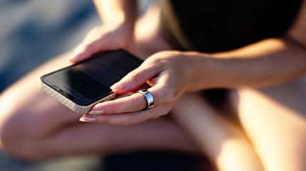Frau mit Smart Ring sitzt am Strand und ihr Smartphone in der Hand