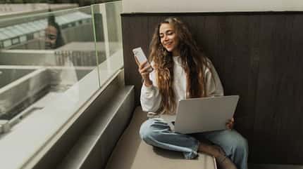Mädchen sitzt auf einer Fensterbank mit Laptop auf dem Schoß und schaut lächelnd auf ihr Smartphone