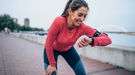 Frau macht draußen eine Pause vom Laufen, stützt sich mit einer Hand auf dem Bein ab und schaut lächelnd auf ihre Fitness Watch