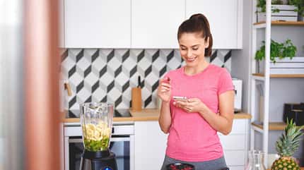 Frau steht in der Küche, bereitet einen Smoothie vor und schaut lächelnd auf ihr Smartphone Frau steht in der Küche, bereitet einen Smoothie vor und schaut lächelnd auf ihr Smartphone