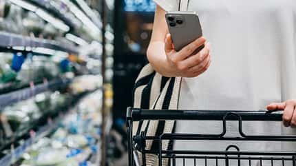 Nahaufnahme: Frau schiebt einen Einkaufswagen im Supermarkt und schaut dabei auf ihr Smartphone Nahaufnahme: Frau schiebt einen Einkaufswagen im Supermarkt und schaut dabei auf ihr Smartphone
