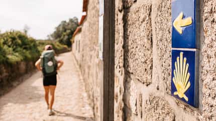 Frau beim Wandern, im Vordergrund ein Wegweiser mit dem Symbol des Jokobswegs Frau beim Wandern, im Vordergrund ein Wegweiser mit dem Symbol des Jokobswegs