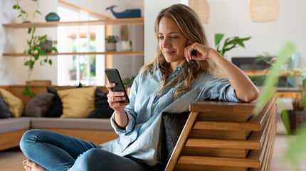 Eine Frau sitzt in ihrem Wohnzimmer im Schneidersitz auf einem Stuhl und schaut auf ihr Smartphone Eine Frau sitzt in ihrem Wohnzimmer im Schneidersitz auf einem Stuhl und schaut auf ihr Smartphone