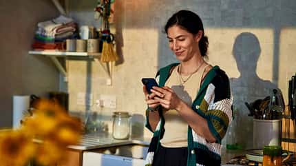 Eine lächelnde Frau steht in der Küche und schaut auf ihr Smartphone Eine lächelnde Frau steht in der Küche und schaut auf ihr Smartphone