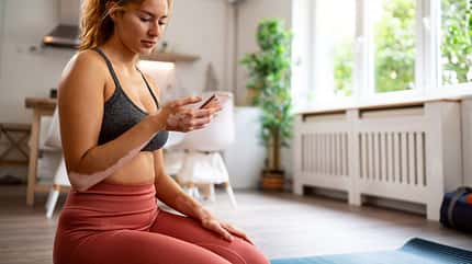 Frau in Sportkleidung sitzt im Wohnzimmer auf einer Yogamatte und schaut auf ihr Handy Frau in Sportkleidung sitzt im Wohnzimmer auf einer Yogamatte und schaut auf ihr Handy