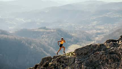 Eine Frau in Laufkleidung läuft einen Bergpass bergauf. Im Hintergrund erstreckt sich ein beeindruckendes Bergpanorama.