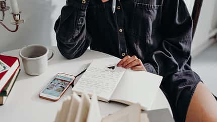 Notizbuch liegt auf Tisch und Frau hält Stift in der Hand.