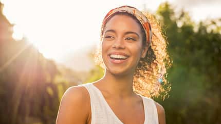 Frau lächelt mit Sonne und Wald im Hintergrund.