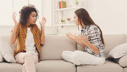 Zwei Frauen sitzen auf einem weißen Sofa und streiten sich. Die linke Frau ist dunkelhäutig, hat braune Locken und trägt ein orangenes Hemd. Die rechte Frau ist weiß, hat braune glatte Haare und trägt ein gemustertes Hemd.