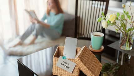 Frau sitzt auf dem Sofa im Hintergrund im Hintergrund. Im Vordergrund steht eine Kiste wo das Handy drin ist, mit einem Zettel dran wo "Digital Detox" draufsteht