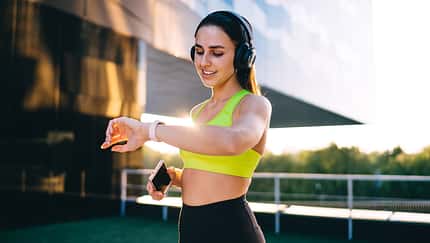 Cheerful caucasian female jogger in headphones enjoying music in headphones and using wearable device on wrist for GPS and time tracking on training, smiling 20s sporty woman looking at smartwatch
