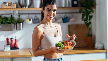 trainierte Frau, die eine gesunde Poke Bowl isst, während sie zu Hause in der Küche in die Kamera schaut