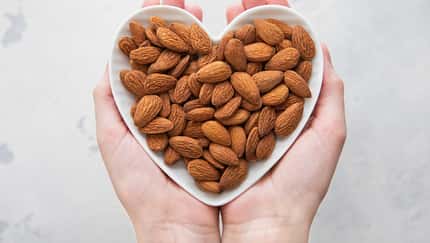 Female hands holding heart shaped plate with peeled almond nuts on light background.Macro.