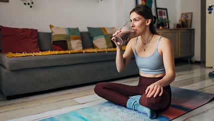 Millennial woman drinks a milkshake while sitting on an exercise mat in the living room