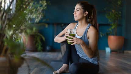 Frau sitzt in Sportkleidung auf einer Yoga-Matte und trinkt grünen Tee