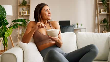 Junge Frau sitzt in Trainingskleidung auf dem Sofa und isst Oatmeal