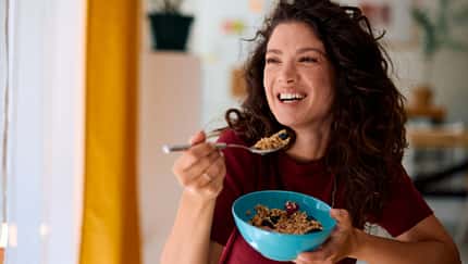 Eine Frau isst lachend ein gesundes Müsli aus einer blauen Schale 