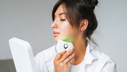 Brunette young woman in white home clothes with clean fresh face doing fasial massage with microcurrent facial massager, home body care