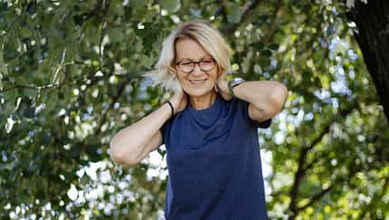 Eine lächelnde ältere Frau mit blonden Haaren und Brille steht im Freien unter grünen Bäumen, trägt ein dunkelblaues T-Shirt und legt entspannt beide Hände in den Nacken.