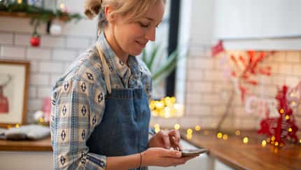Frau mit Schürze steht in einer weihnachtlich geschmückten Küche und schaut auf ihr Smartphone
