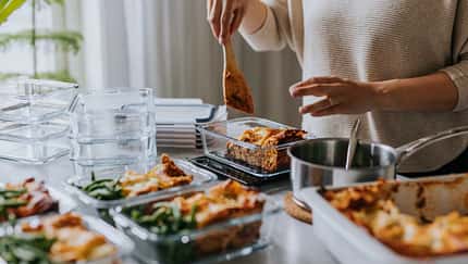 Eine Frau befüllt mehrere Brotdosen mit Lasagne. Die Arbeitsfläche ist gefüllt mit leeren und befüllten Brotdosen