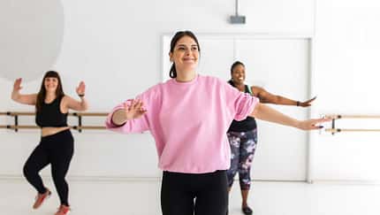 Drei Frauen machen gemeinsam ein Tanz-Workout in einem hellen Fitnessstudio, die Frau vorne trägt ein rosa Shirt und lächelt.