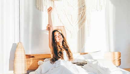 Junge Frau mit langen Haaren sitzt zugedeckt im Bett und streckt sich genüsslich Junge Frau mit langen Haaren sitzt zugedeckt im Bett und streckt sich genüsslich