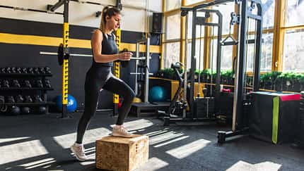 Frau macht Step-Up Übung in Fitnessstudio, im Hintergrund mehrere Fitnessgeräte