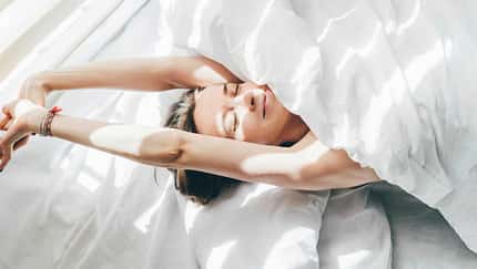 Frau liegt unter einer weißen Decke im Bett und streckt sich genüsslich Frau liegt unter einer weißen Decke im Bett und streckt sich genüsslich