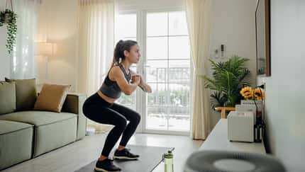 Frau in Sportkleidung macht Kniebeugen auf einer Matte im Wohnzimmer vor einer Terrassentür, neben Sofa und Zimmerpflanze.