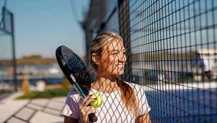 Frau mit Padelschläger und Ball steht lächelnd an einem Metallzaun, Sonnenlicht wirft Gittermuster auf ihr Gesicht.