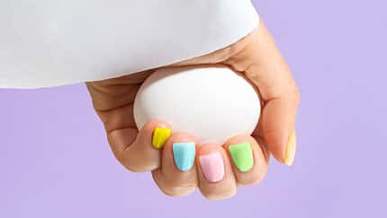 Elegant woman's hand with pastel color manicure holding a white egg on Easter Holiday on Trendy Color of the Year 2022 Very Peri purple violet lavender color background. Woman dressed in fashionable white shirt.