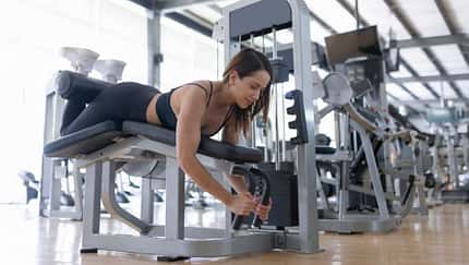 Frau trainiert im Fitnessstudio an einer Beinbeuger-Maschine in Bauchlage und führt eine kontrollierte Kraftübung aus.