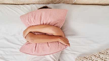 Eine Frau liegt im Bett und verbirgt ihr Gesicht hinter einem Kissen, das sie mit beiden Armen umschlungen hält Eine Frau liegt im Bett und verbirgt ihr Gesicht hinter einem Kissen, das sie mit beiden Armen umschlungen hält