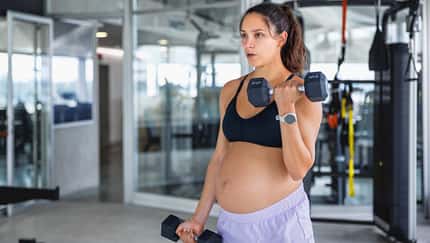 Schwangere Frau steht in einem Fitnessstudio und trainiert mit zwei Hanteln Schwangere Frau steht in einem Fitnessstudio und trainiert mit zwei Hanteln