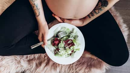 Eine schwangere Frau im Schneidersitz streichelt ihren Bauch und hat einen Teller mit Salat auf dem Schoß