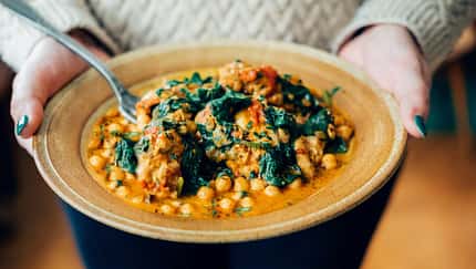 Zwei Frauenhände halten eine Schale mit einem Kichererbsen-Spinat-Curry Zwei Frauenhände halten eine Schale mit einem Kichererbsen-Spinat-Curry