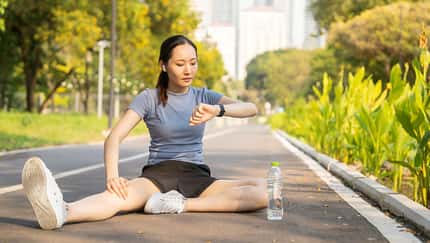 Eine Frau sitzt auf der Straße und stretcht sich in Sportkleidung während sie auf ihre Fitnessuhr schaut